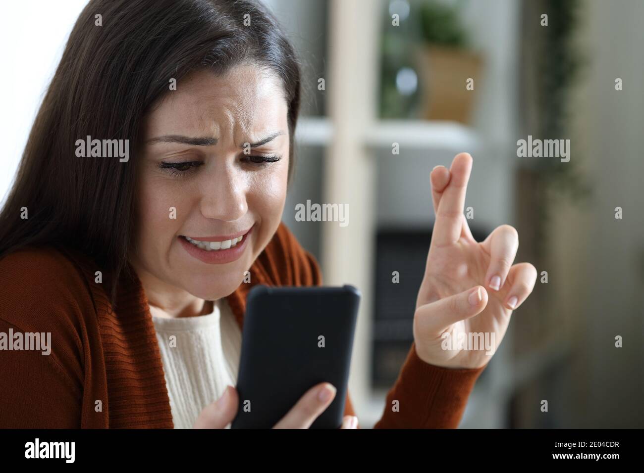 Nervous woman crossing fingers waiting news on mobile phone at home Stock Photo