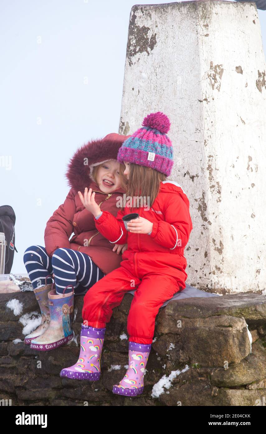 Two young girls sat talking sitting on the trig point or triangulation point on the summit of Bosley Cloud near Congleton Cheshire  in the winter snow Stock Photo