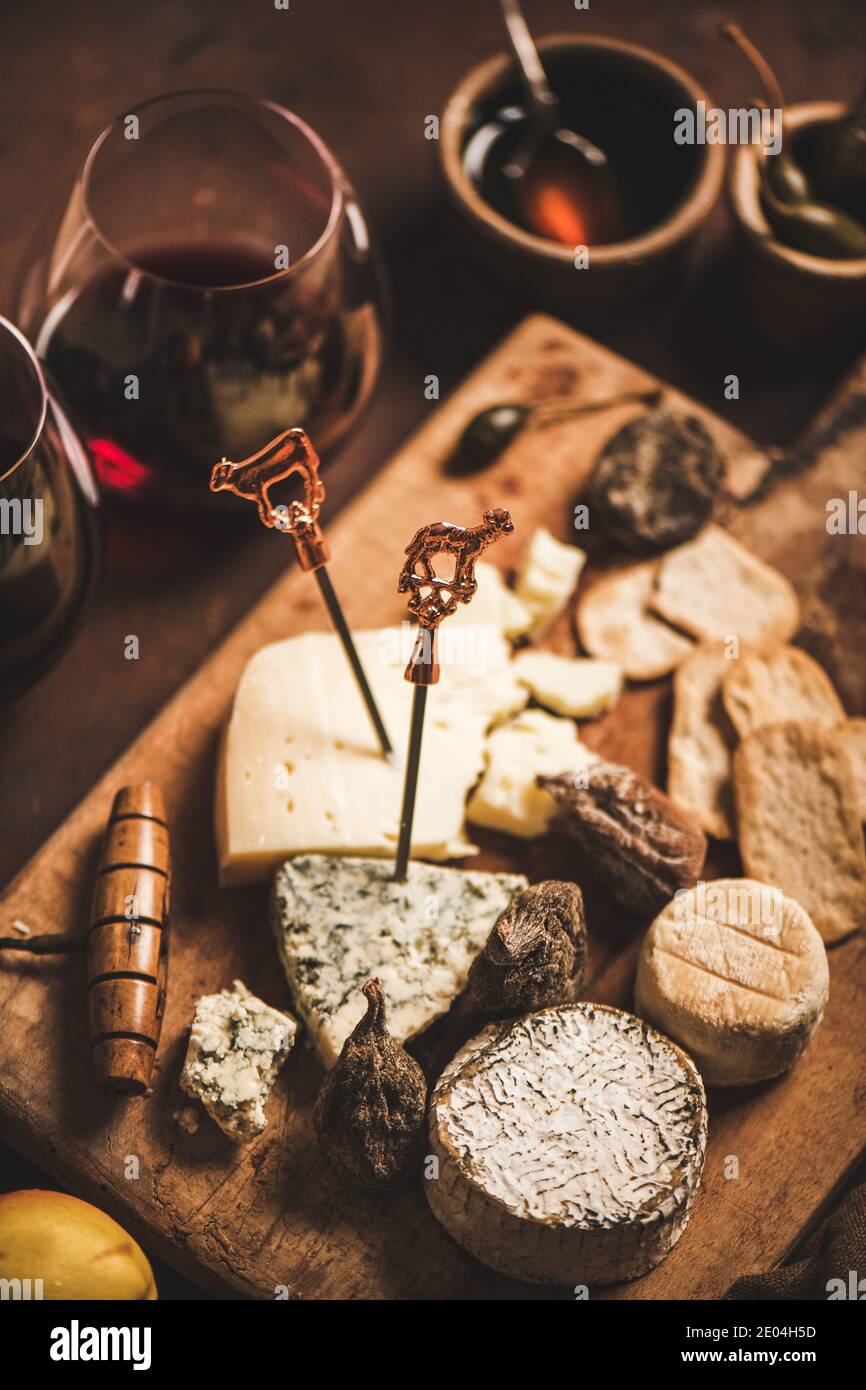 Assortment of snacks and appetizers for red wine. Various cheeses on sticks with bull, crackers, dry fruit and glasses of red wine on wooden rustic board over rusty brown background, selective focus Stock Photo