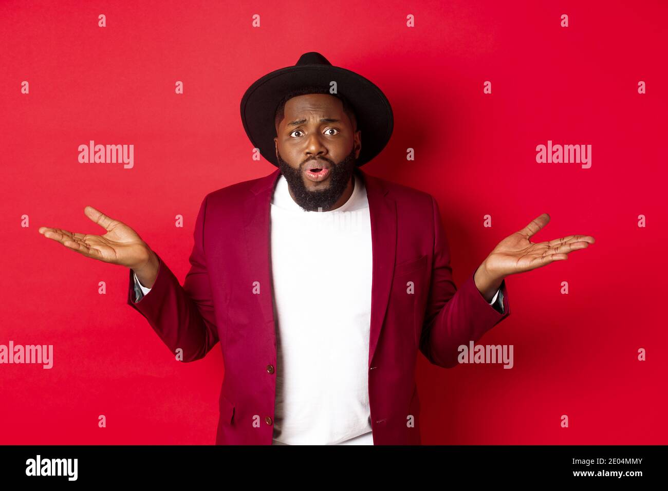 Confused and worried Black man spread hands sideways, staring at camera puzzled, asking what wrong, standing against red background Stock Photo