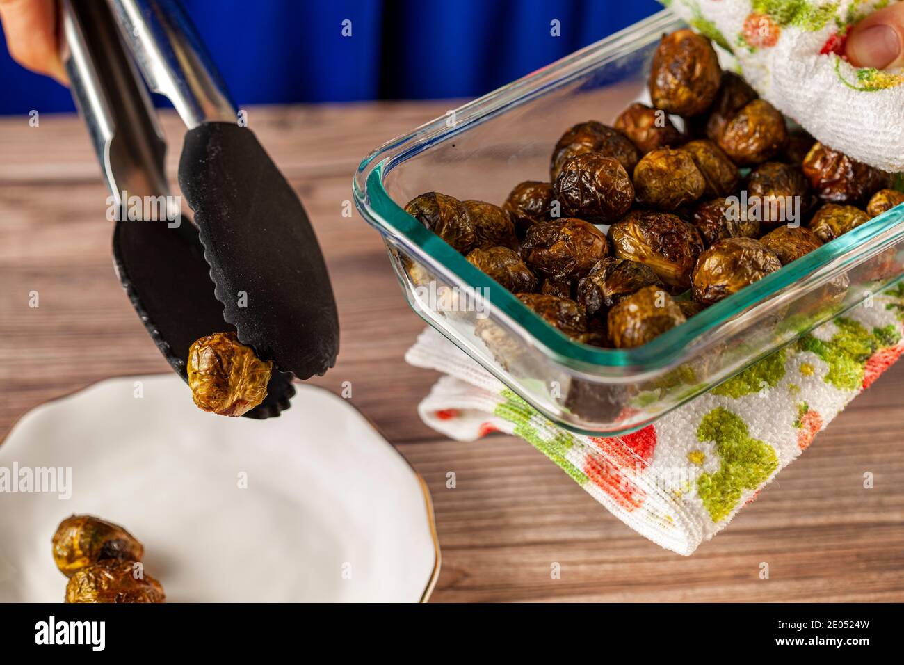 Close up isolated image of a thick glass bowl of cooked and seasoned Brussel sprouts being held with kitchen cloth and served into porcelain plate on Stock Photo