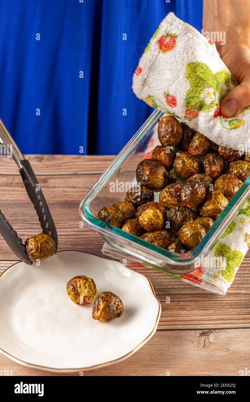 Close up image of a thick glass bowl of freshly cooked and seasoned Brussel sprouts being hold by a caucasian woman with kitchen cloth and served into Stock Photo