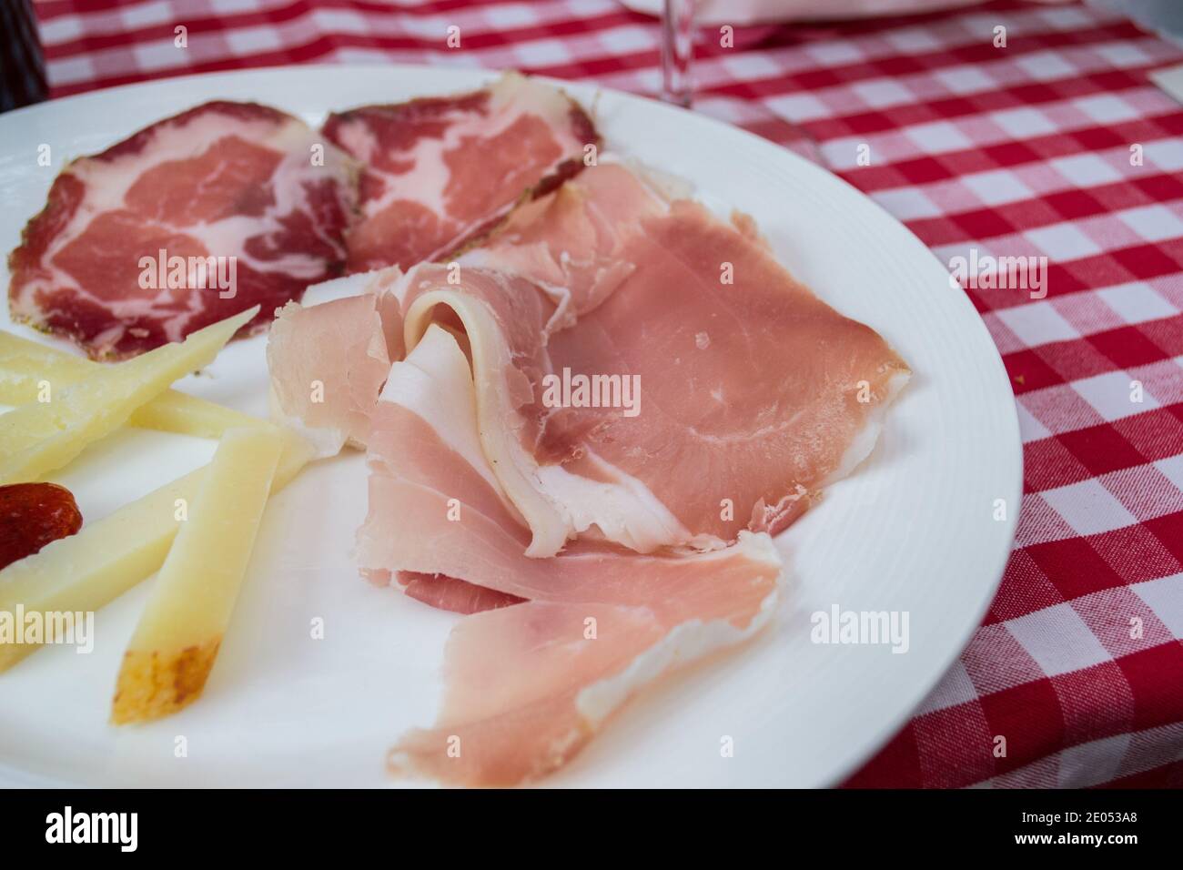 White plate on red and white tablecloth, cheese, sliced meats, appetizer snack plate, picnic, prosciutto Stock Photo