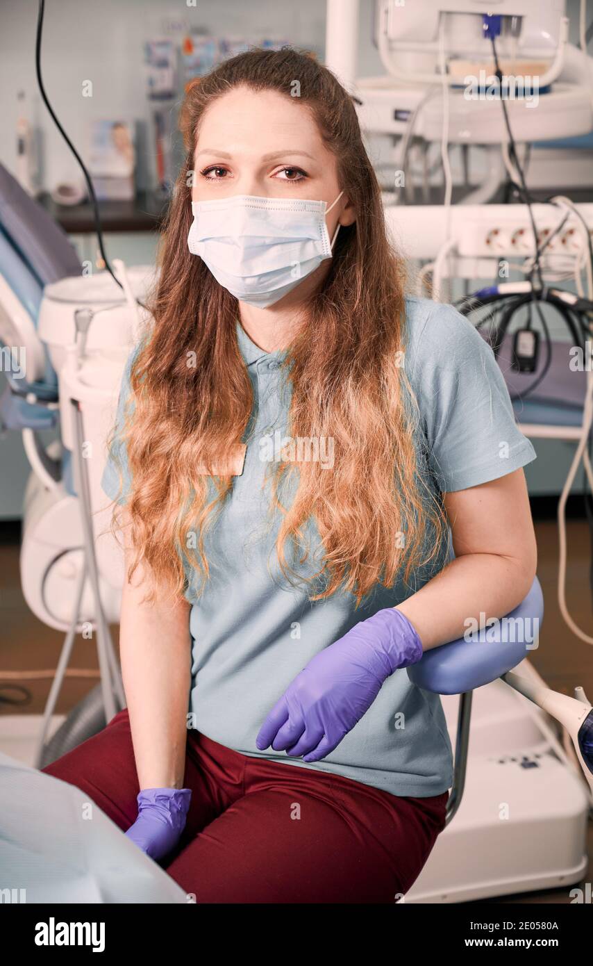 Portrait of charming female dentist in medical mask with long red hair posing in dental office