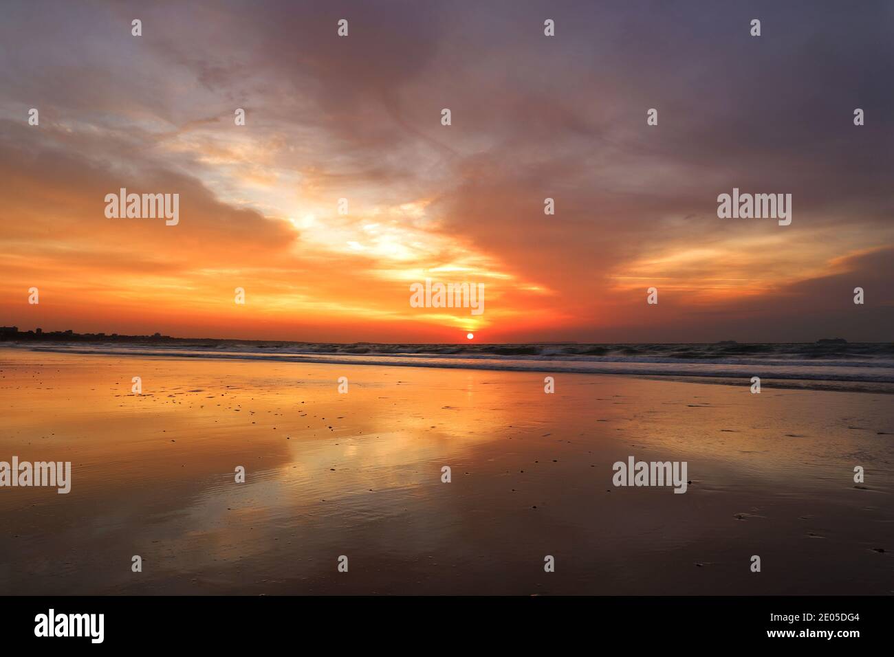 The sun rises in a blaze of fiery orange and red colours into a cloudy winter sky, all reflected in the wet sand of Bournemouth beach. Stock Photo