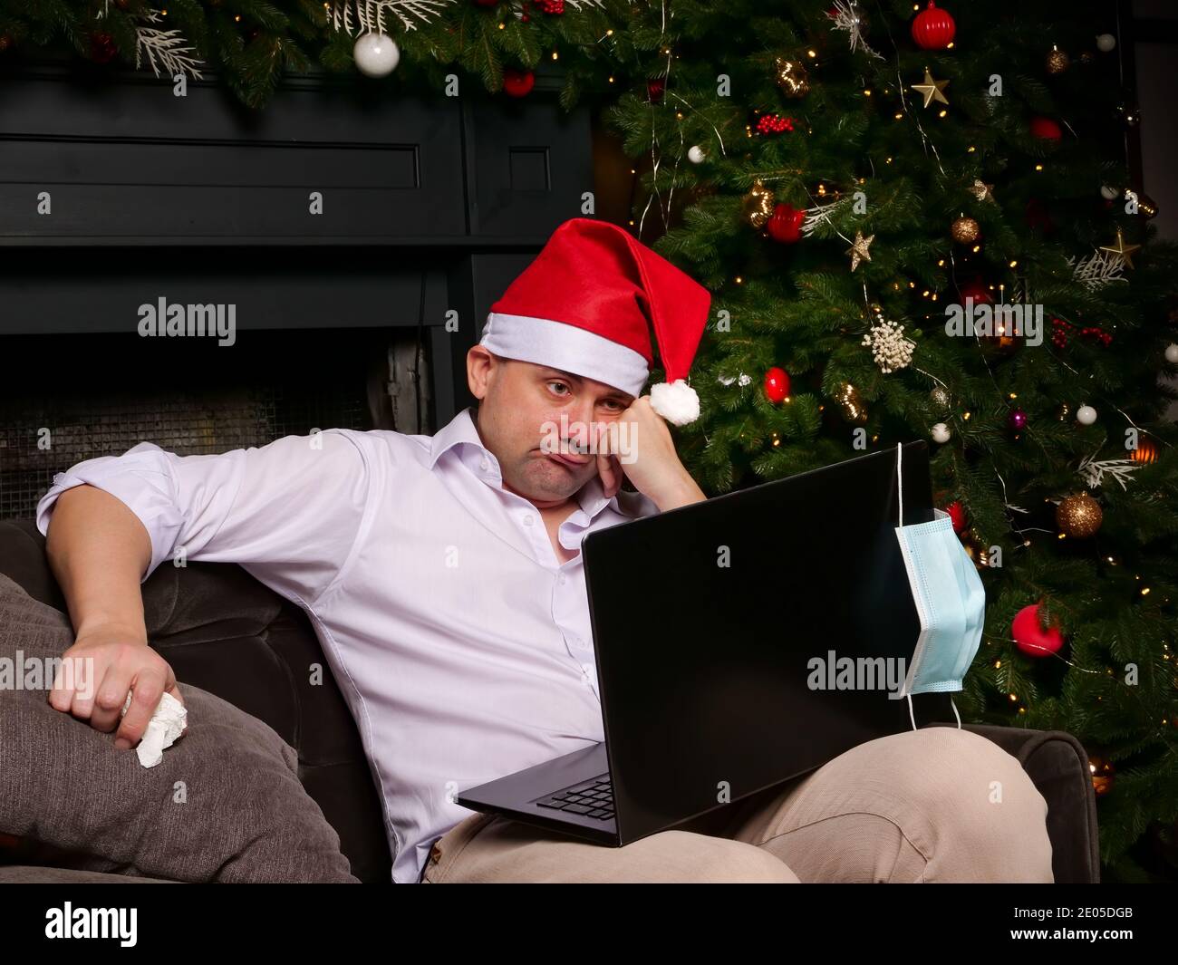A sad sick man in a Santa hat is looking at a laptop alone. New Years celebration and covid epidemic. Stock Photo