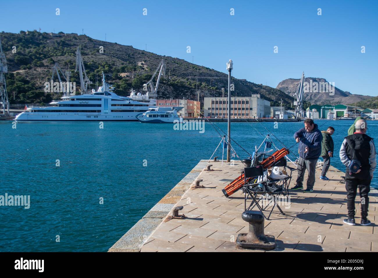 Fishemen on the quayside as a tourist boat sailing past the yacht 'Vibrant Curiosity' moared in the marina at Cartagena, Murcia, Spain Stock Photo