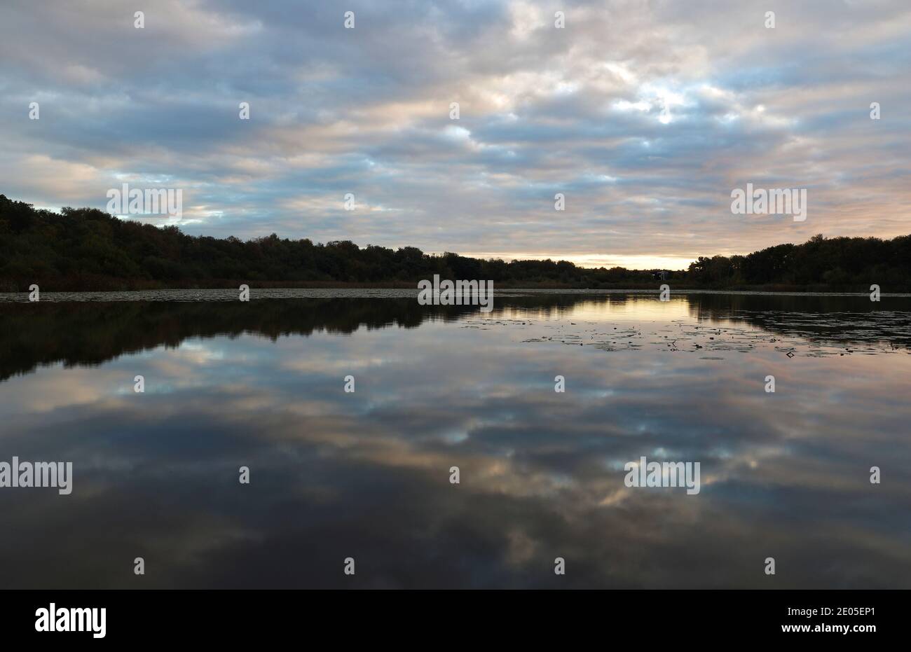 The rising sun sprays an arch of light into the cloud cover above the glass-like water of Hatch Pond. Trees line the horizon of this peaceful scene. Stock Photo
