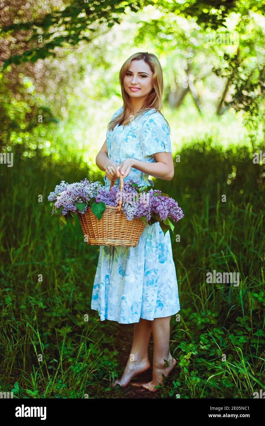 Portrait of young woman in blue dress in spring garden with a basket full of lilacs. Spring background. Woman's Day. Greeting card. Stock Photo