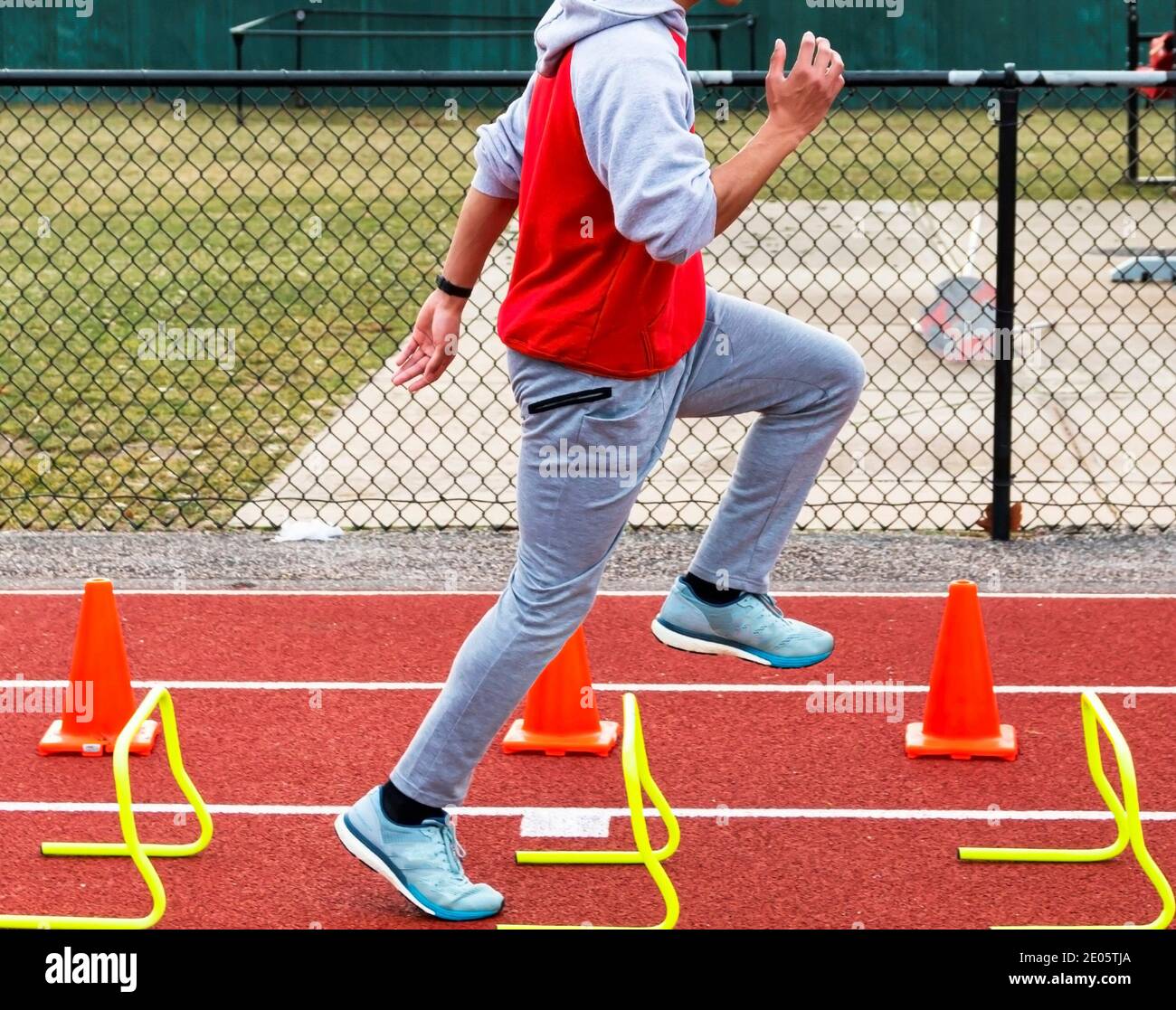 Side view of a high school runner running over yellow mini hurdles and orange construction cones on a red track during track and field practice. Stock Photo