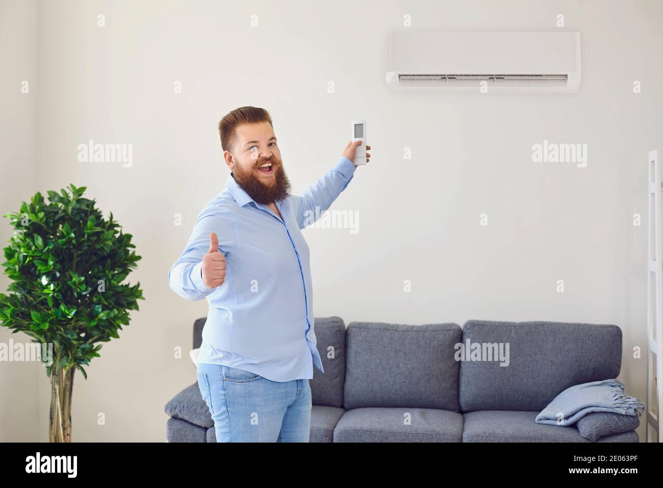 Funny bearded man turning on air conditioner with remote control and showing thumb up in apartment, copy space Stock Photo