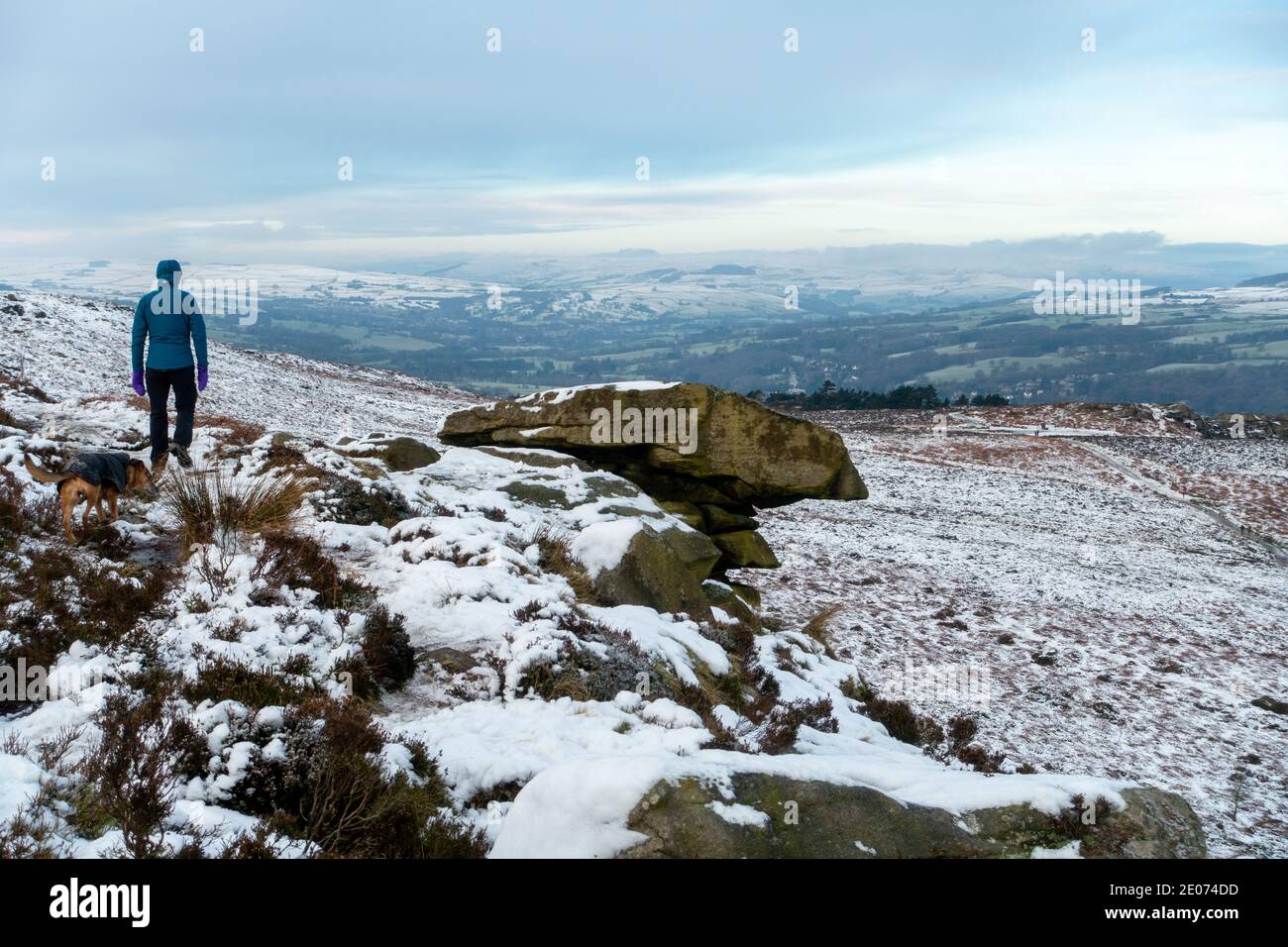 Walker walking a dog next to The Pancake Stone on Ilkley Moor on a snow covered moorland loking towards the Cow and Calf Rocks in West Yorkshire, UK Stock Photo