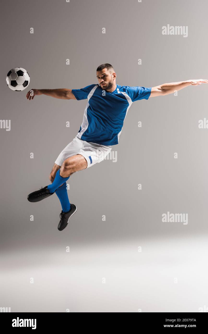 Athletic man jumping with crossed legs near football on grey background Stock Photo
