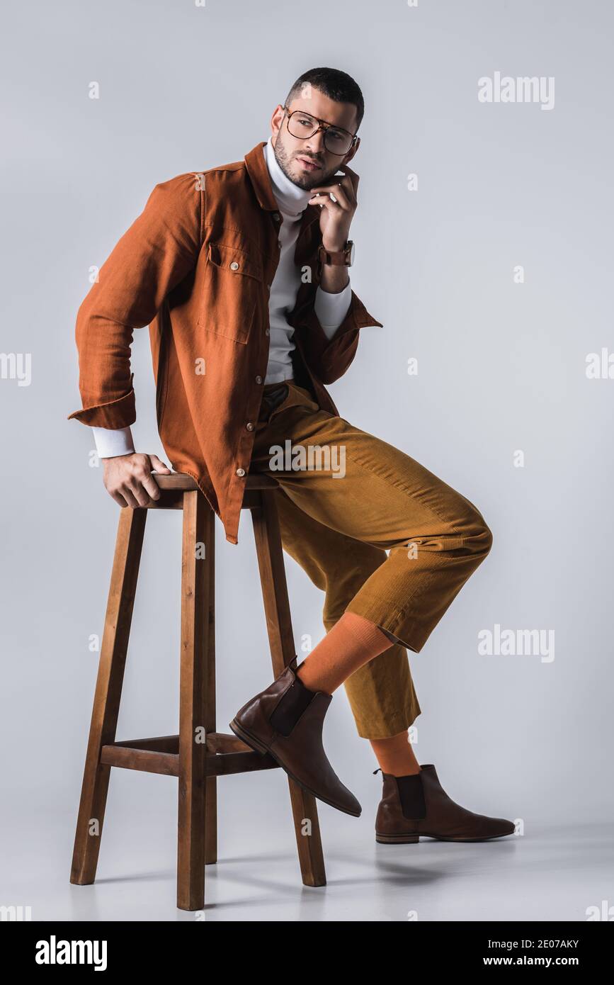 Fashionable man in terracotta jacket and brown shoes posing near wooden chair on grey background Stock Photo