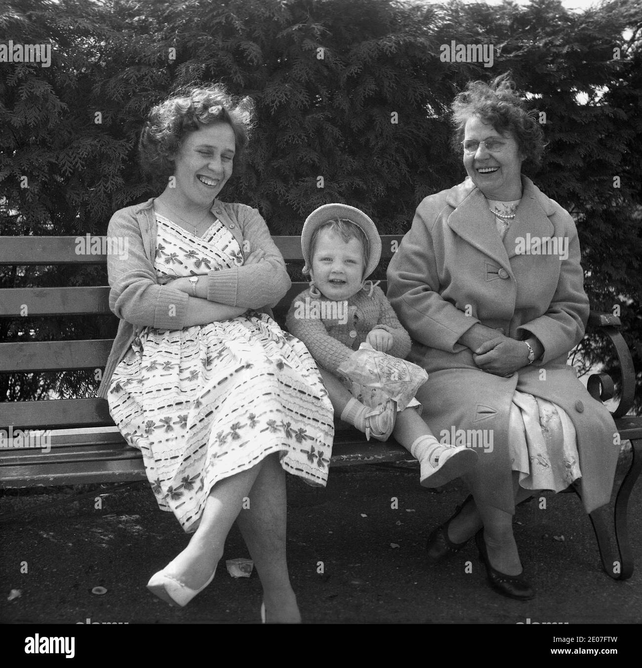 1950s, historical, happy family times, a mother sitting on a bench laughing together with her daughter, who is wearing a bonnet and whose hand is in a crisp packet, alongside the child's grandmother, who had a big smile on her face, England, UK. Stock Photo