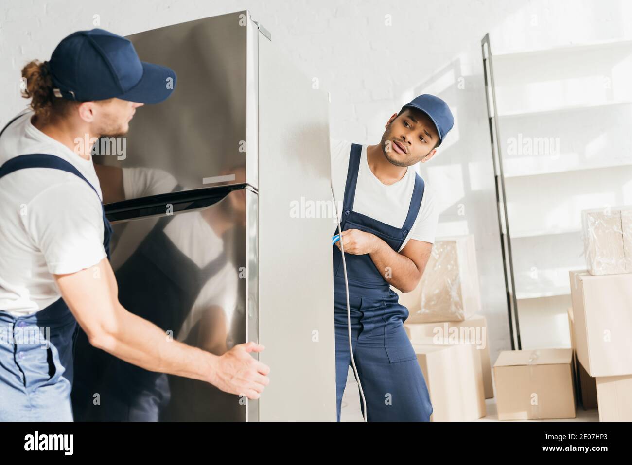 Indian mover looking at coworker while moving fridge in apartment Stock Photo