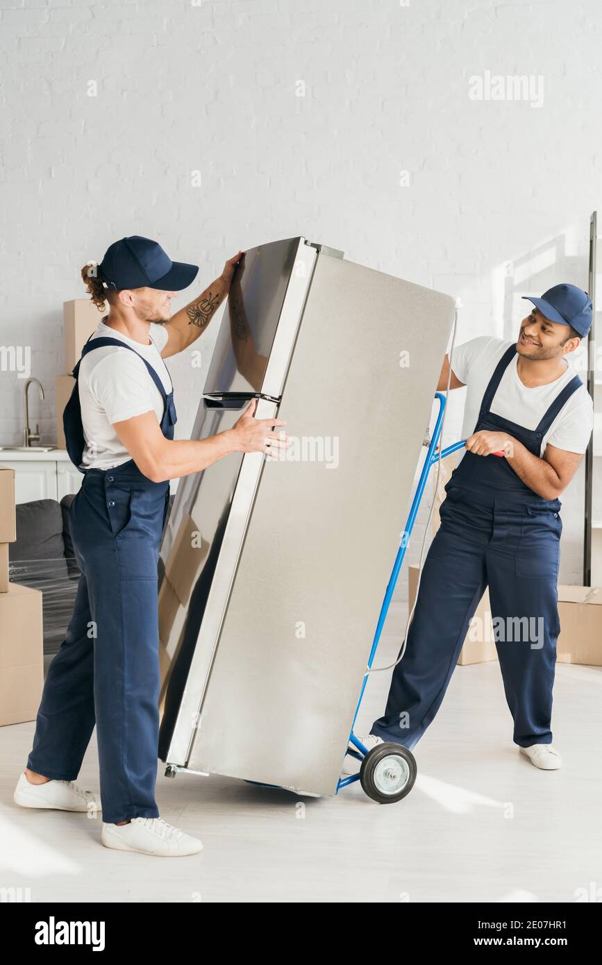cheerful indian mover looking at coworker while moving fridge in apartment Stock Photo