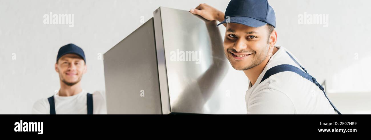 cheerful indian mover in uniform moving fridge near coworker on blurred background, banner Stock Photo