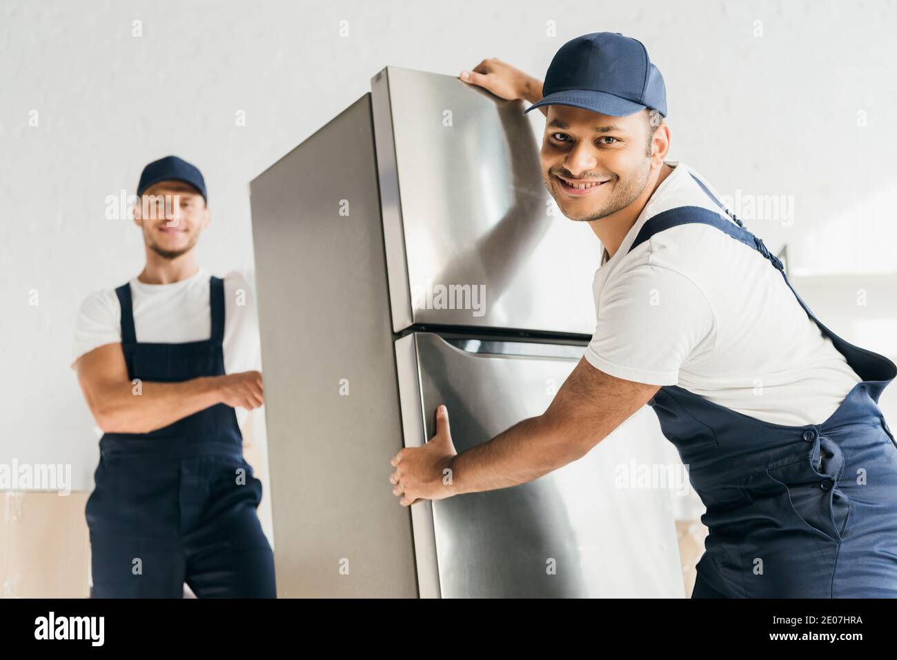 cheerful indian mover in uniform moving fridge near coworker on blurred background Stock Photo