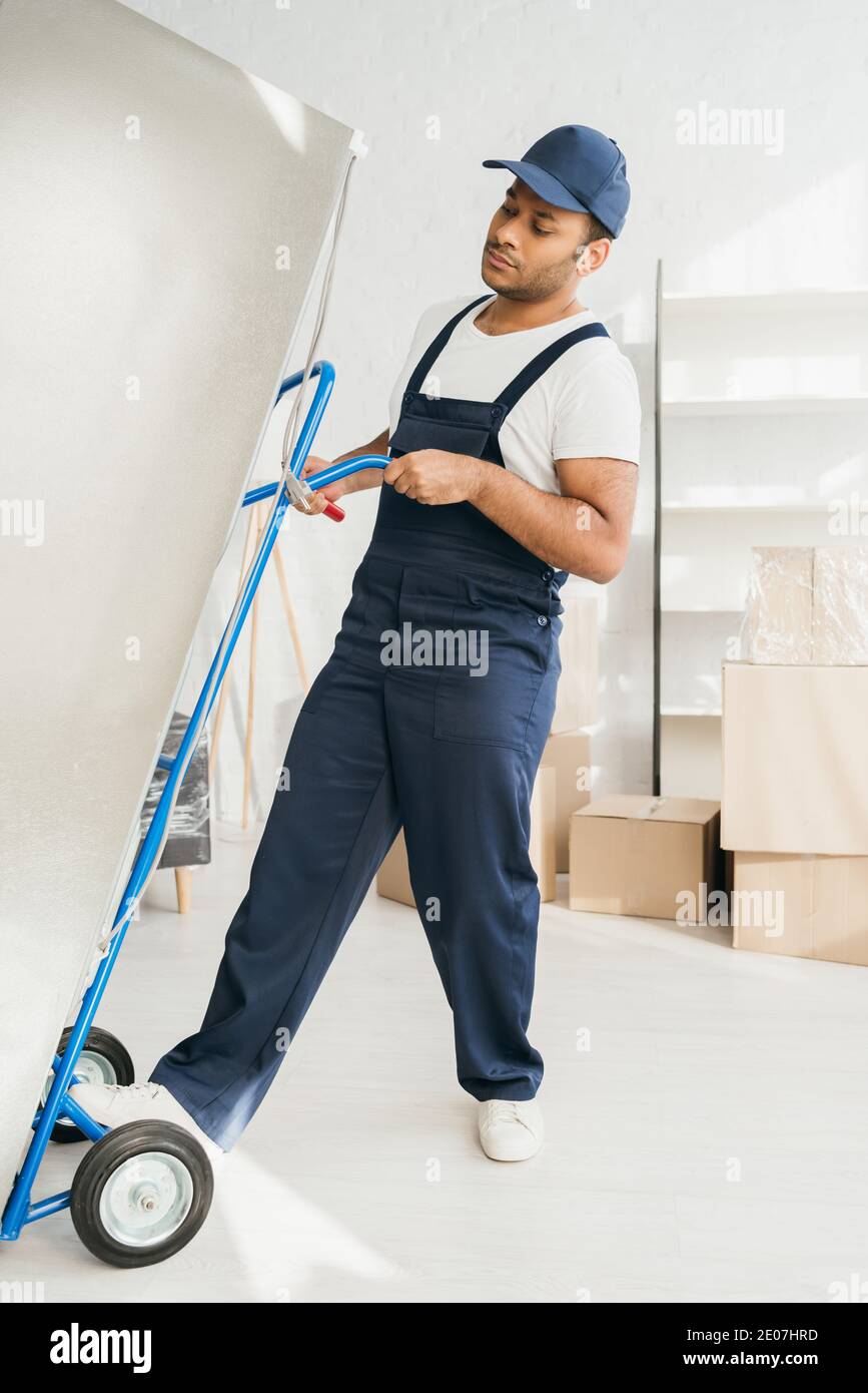 strong indian workman holding hand truck while moving fridge in apartment Stock Photo
