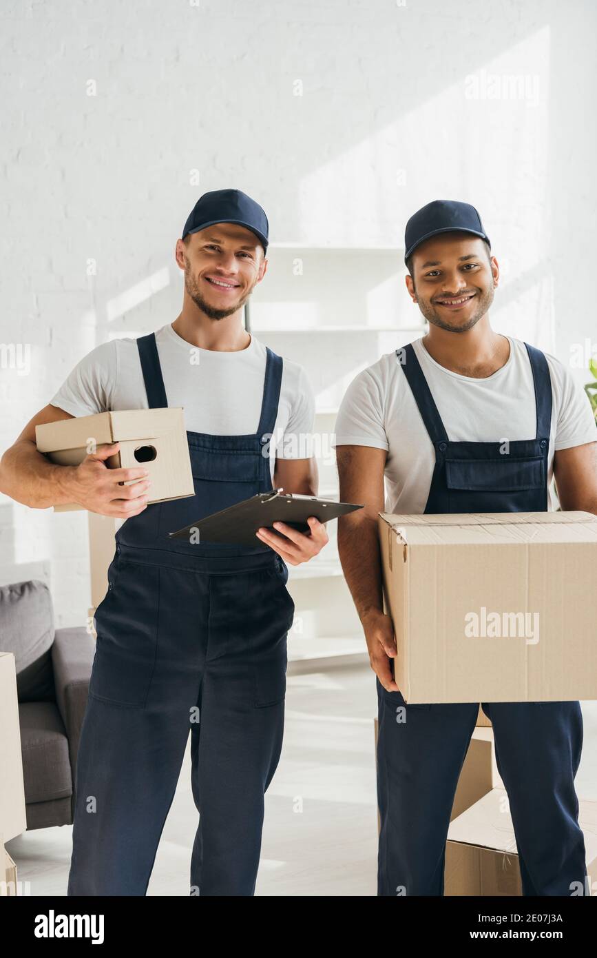 cheerful multicultural movers smiling while looking at camera and holding boxes in apartment Stock Photo