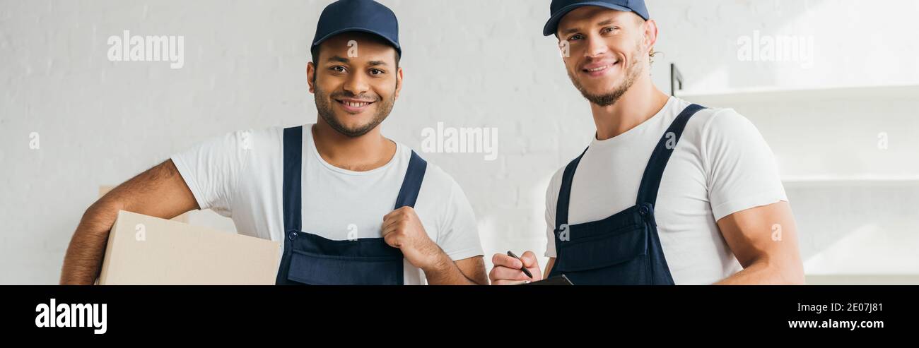 cheerful multicultural movers smiling while looking at camera in apartment, banner Stock Photo