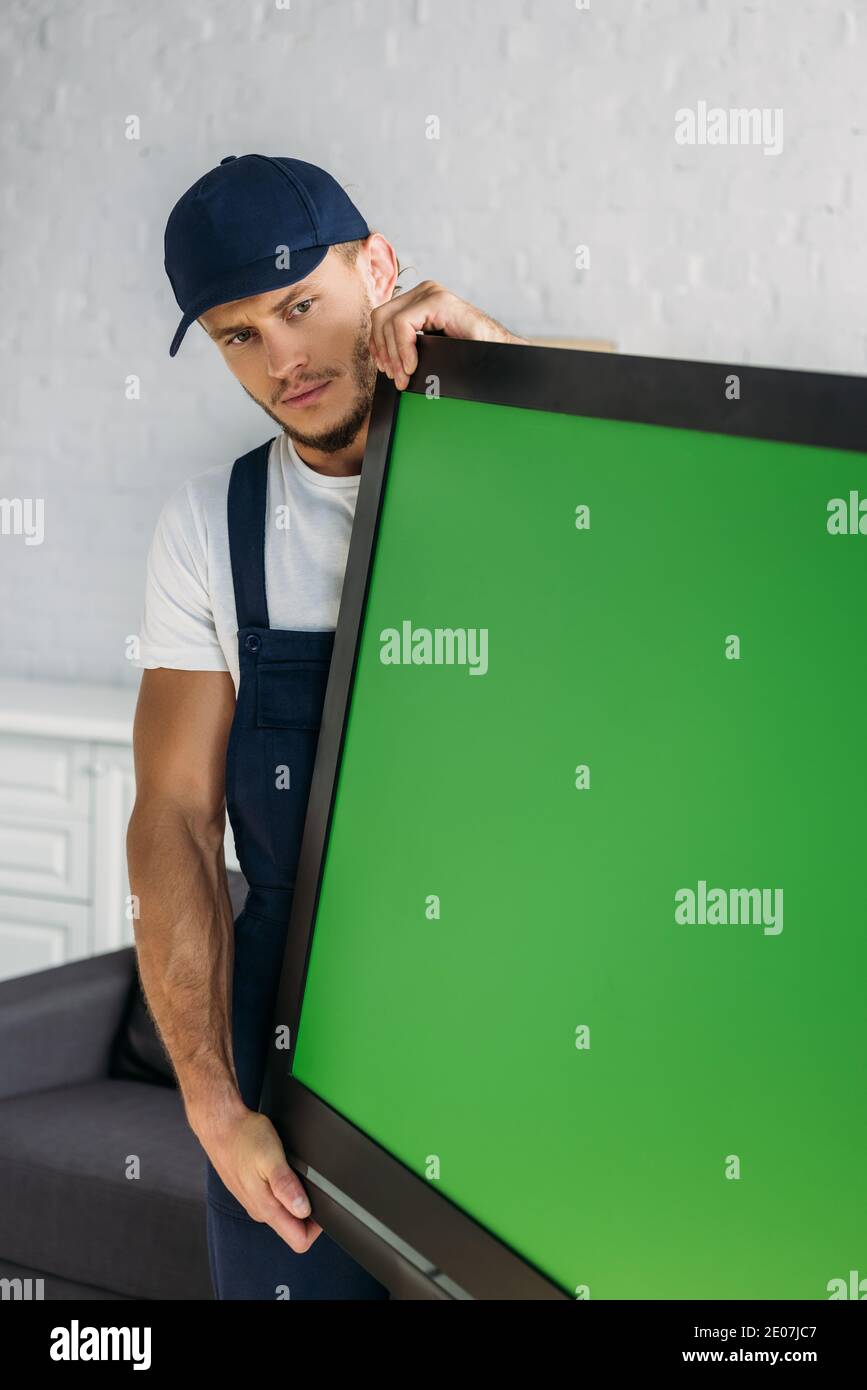 young mover in uniform carrying plasma tv with green screen in apartment Stock Photo