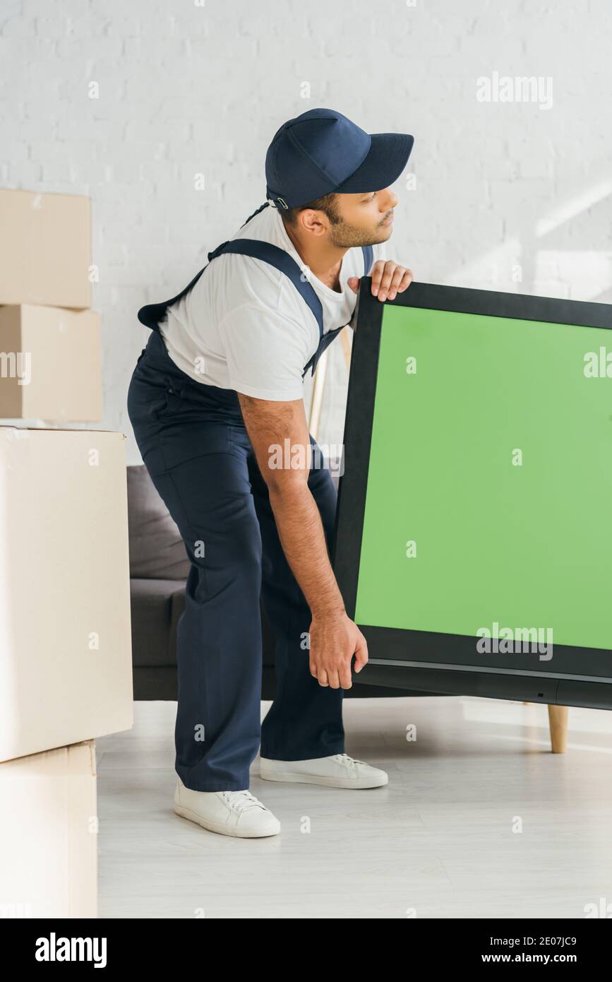 Indian mover in uniform carrying plasma tv with green screen in apartment Stock Photo