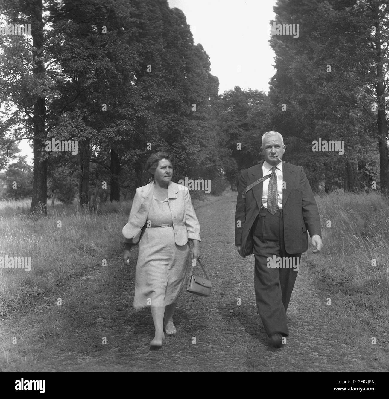 1950s, historical, a middle aged, well-dressed couple walking along a tree-lined grassy pathway in semi-rural parkland, England, UK. The gentleman is wearing a suit and tie and smoking a pipe, while the lady is carrying her handbag, perhaps they are taking a stroll after attending church or a civic function. Stock Photo