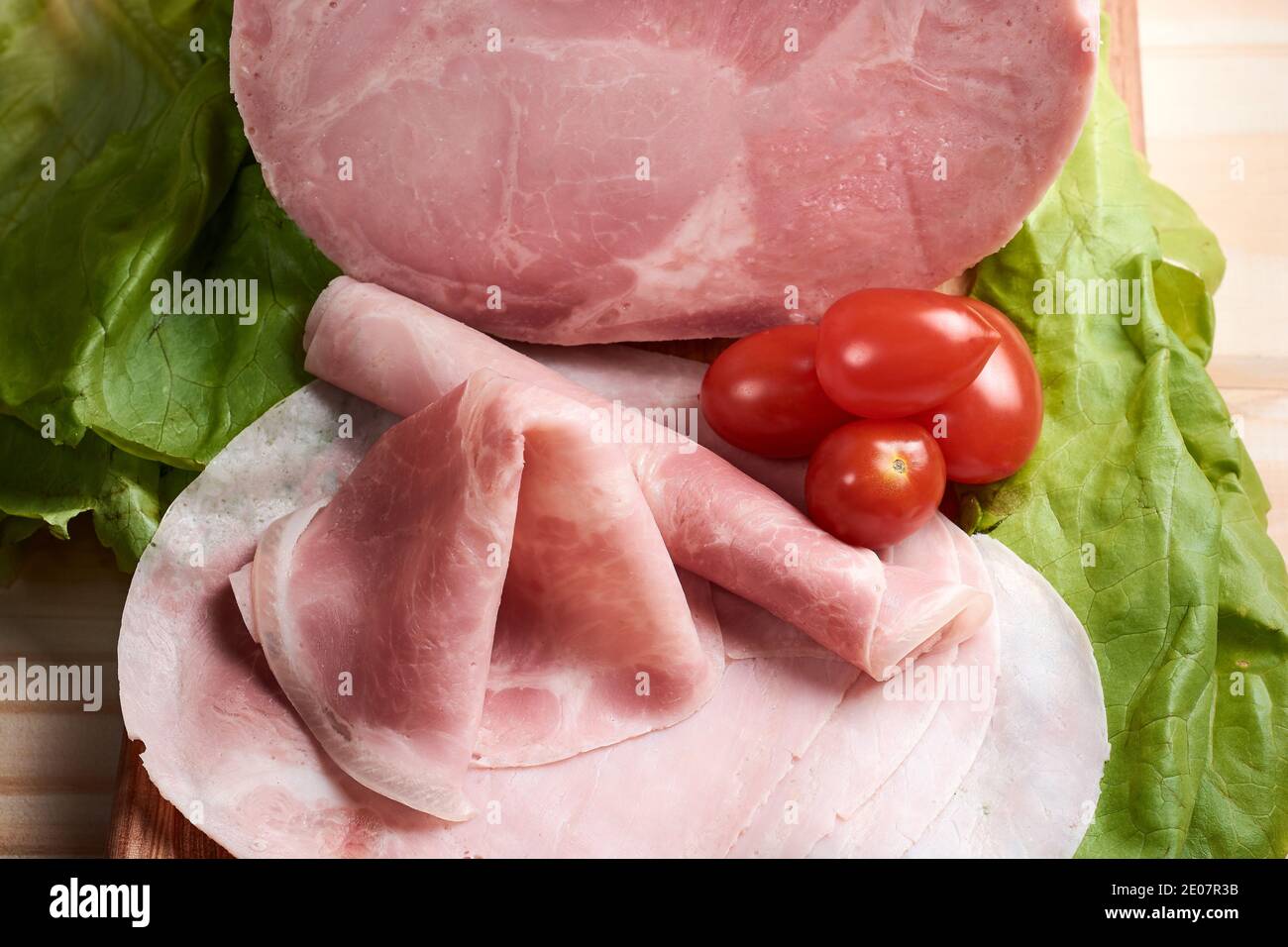 Delicious sliced and rolled cooked pork ham on wooden table accompanied by fresh cherry tomatoes and lettuce. Stock Photo