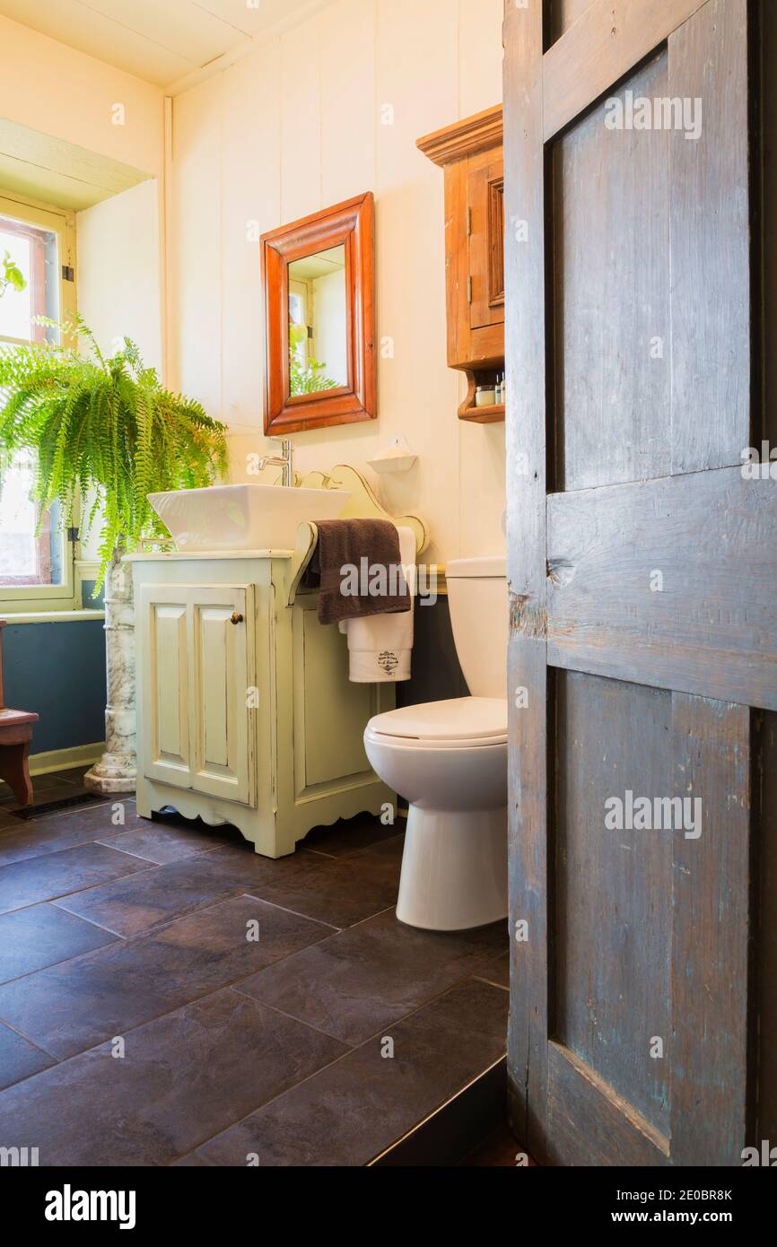 Opened door of main bathroom with ceramic tile floor and white toilet and sink on top of antique cabinet inside an old 1841 cottage style home Stock Photo