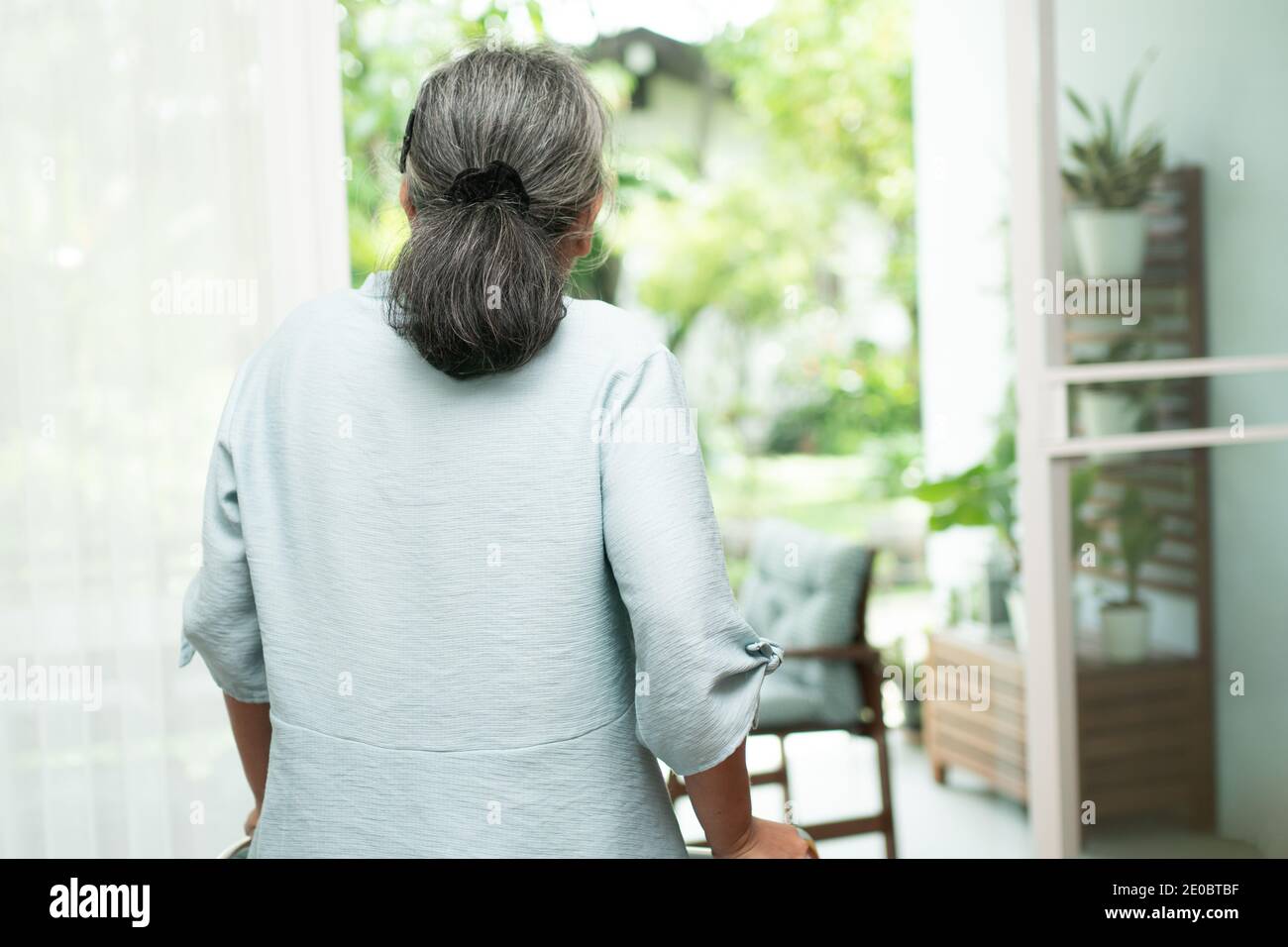 A sad old elderly woman uses Walker for standing in front of windows and looking outside and feeling lonely. Concept of depression caused by illness a Stock Photo