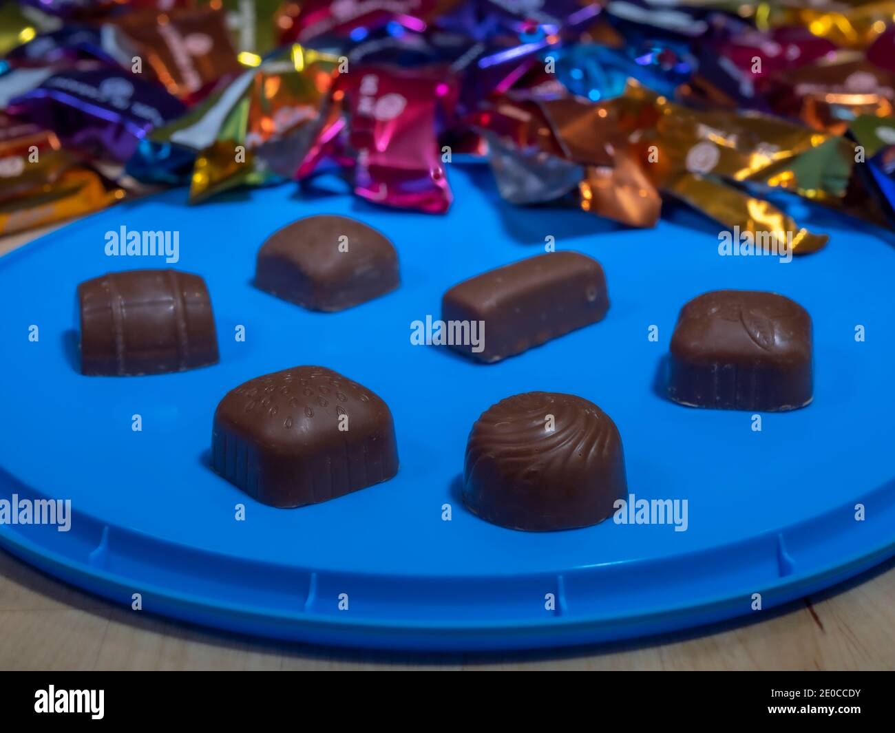 Closeup of filled, coated milk chocolates, with out of focus colourful wrappers in the background, and some in focus opened chocolates on the box lid. Stock Photo