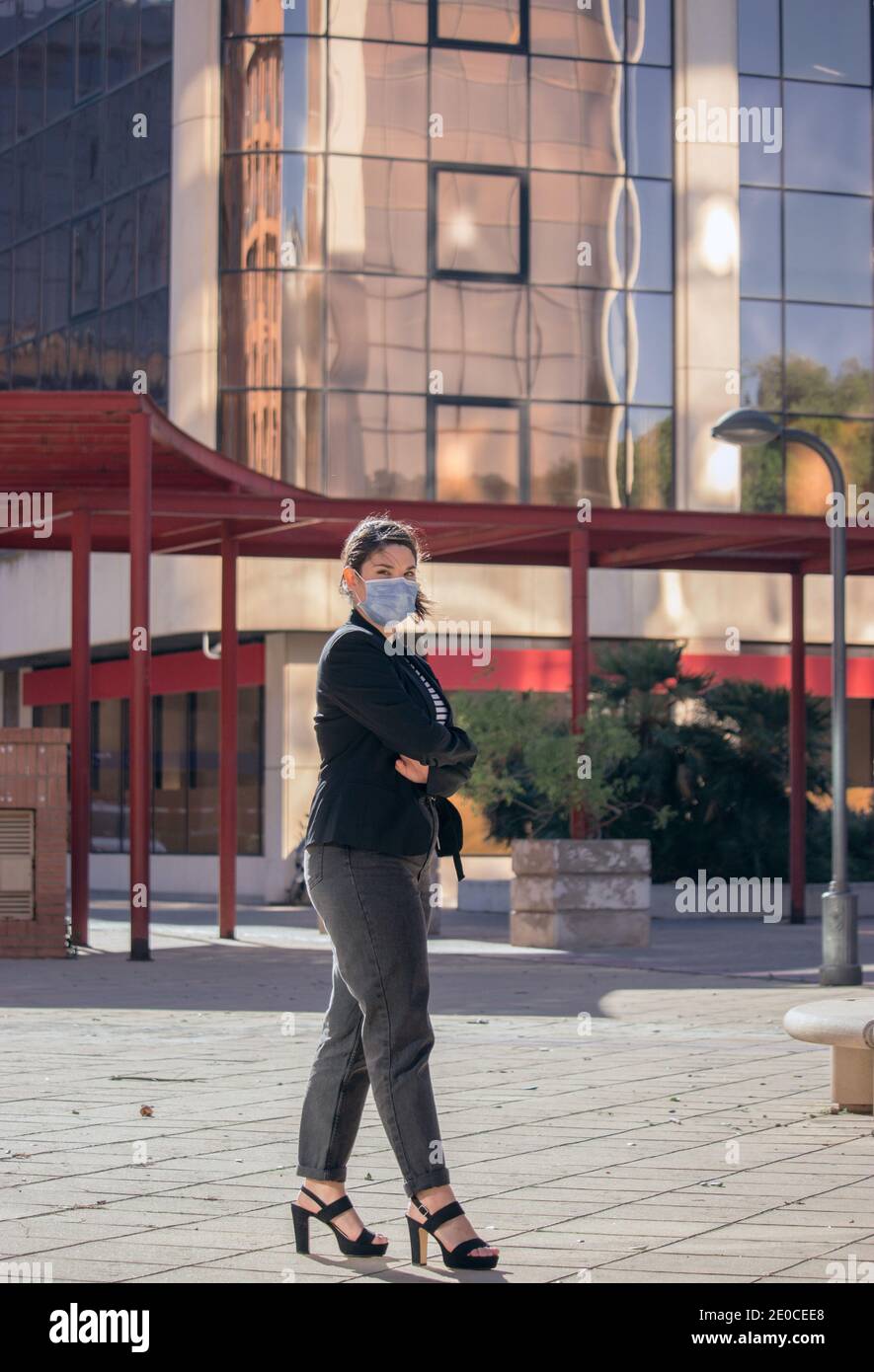Elegant businesswoman with mask on, standing outside the office poses looking straight to the camera outdoor. Girl Boss Concept 2021. Stock Photo