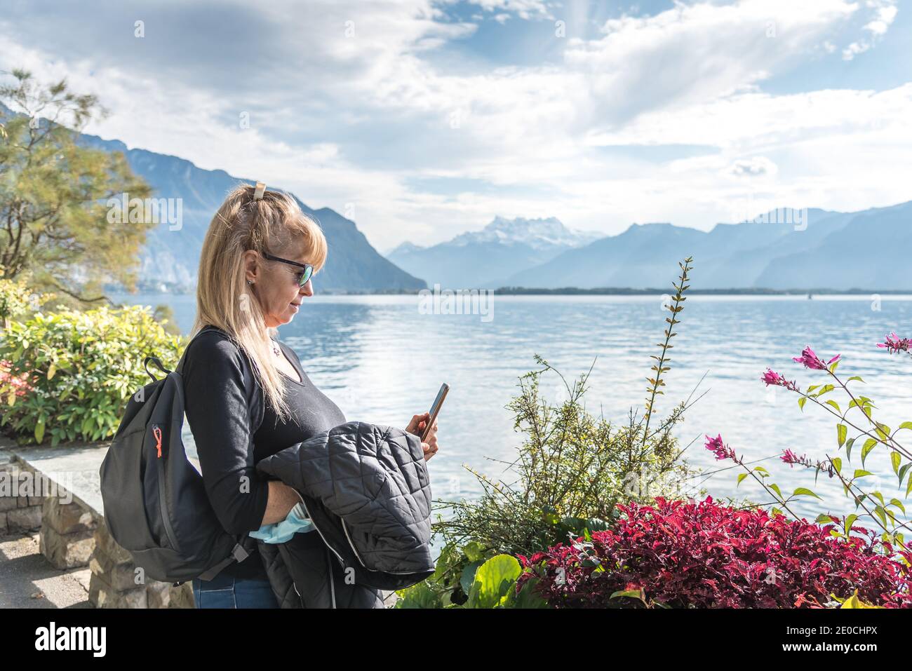 Blonde woman with sunglasses and a bag using her mobile with a mask hanging from his arm in front of a lake surrounded by mountains. Montreux in Genev Stock Photo