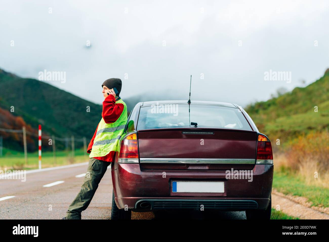 Side view of bewildered female driver in reflective vest leaning on broken car while standing on road in highlands and calling insurance for help Stock Photo