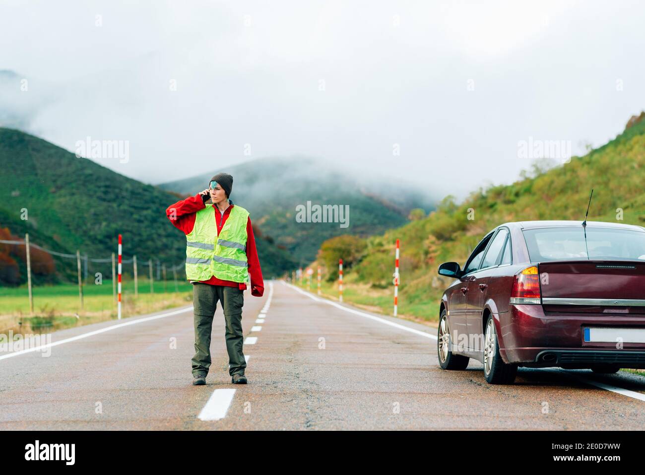 Female driver in reflective vest standing on road near broken car in mountainous area and talking on smartphone while calling for help Stock Photo