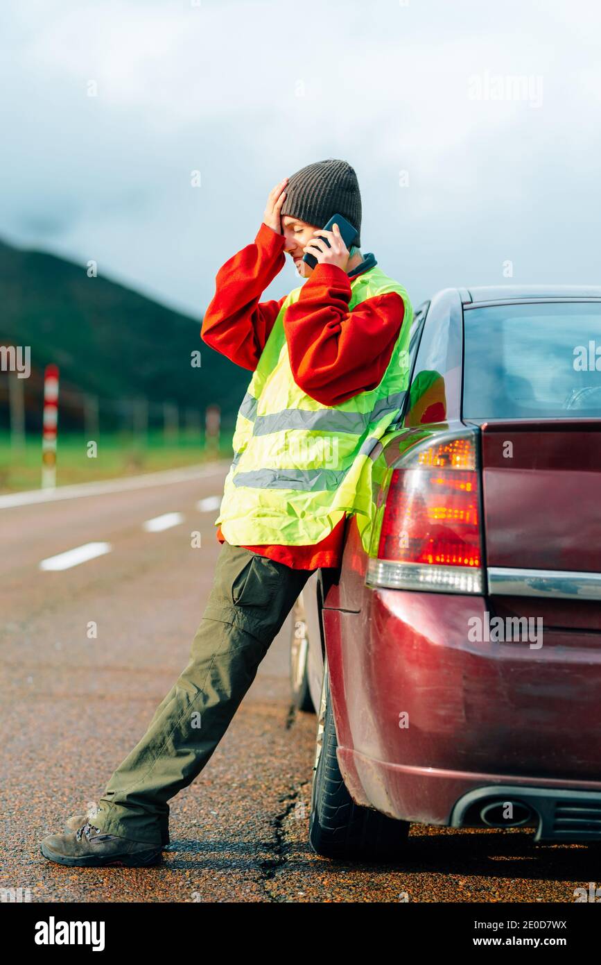 Side view of bewildered female driver in reflective vest leaning on broken car while standing on road in highlands and calling insurance for help Stock Photo