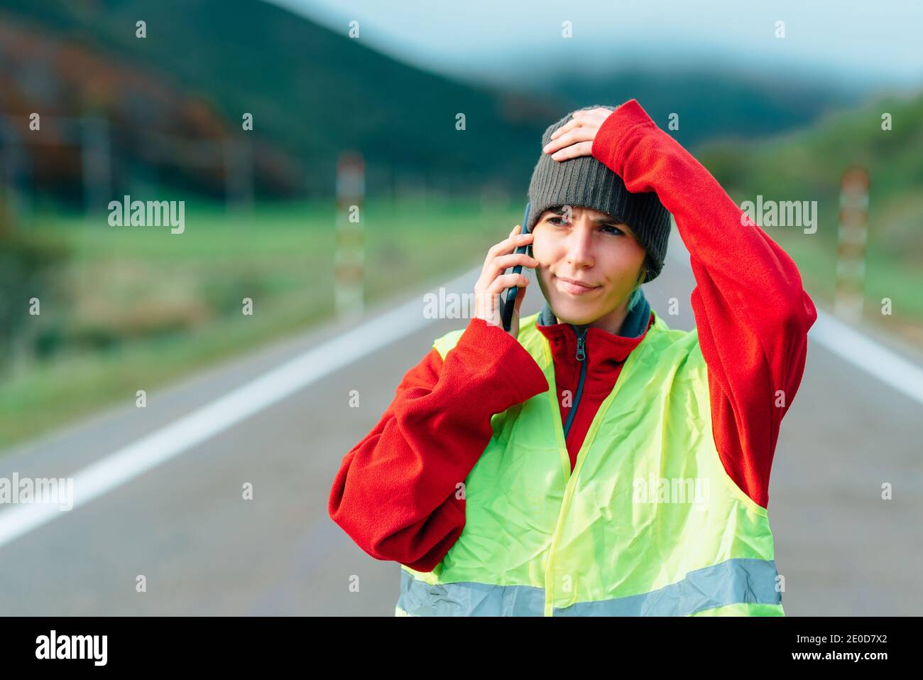 Concerned female driver in reflective vest standing on empty road and talking on smartphone while calling insurance for help Stock Photo