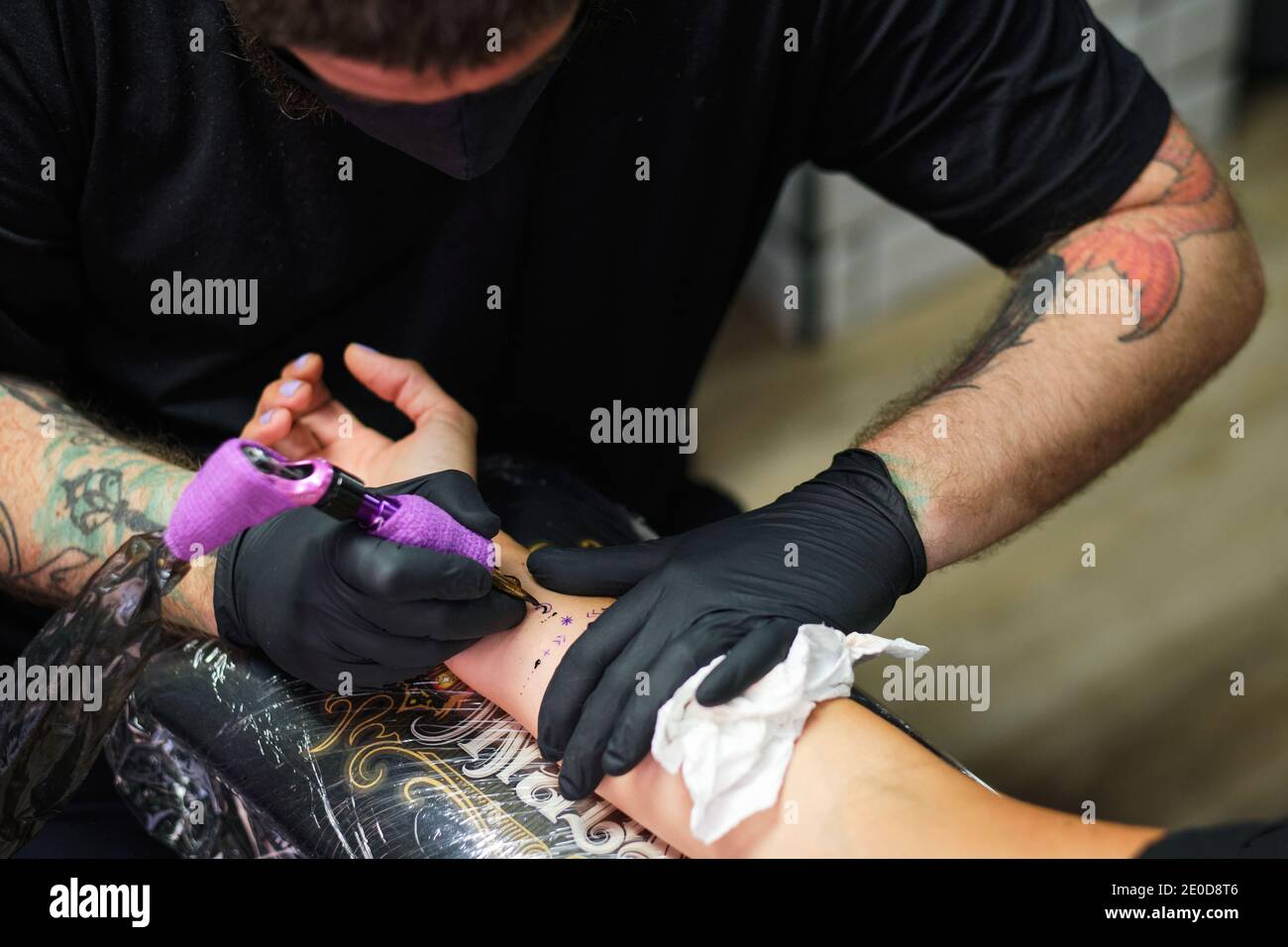 Unrecognizable concentrate bearded male master in black face mask and gloves creating tattoo on arm of crop faceless client in salon Stock Photo