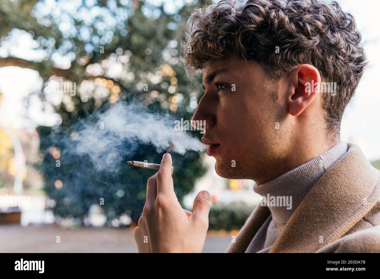 Side view of serious male in stylish outfit standing in street and smoking cigarette while exhaling fume and looking away Stock Photo