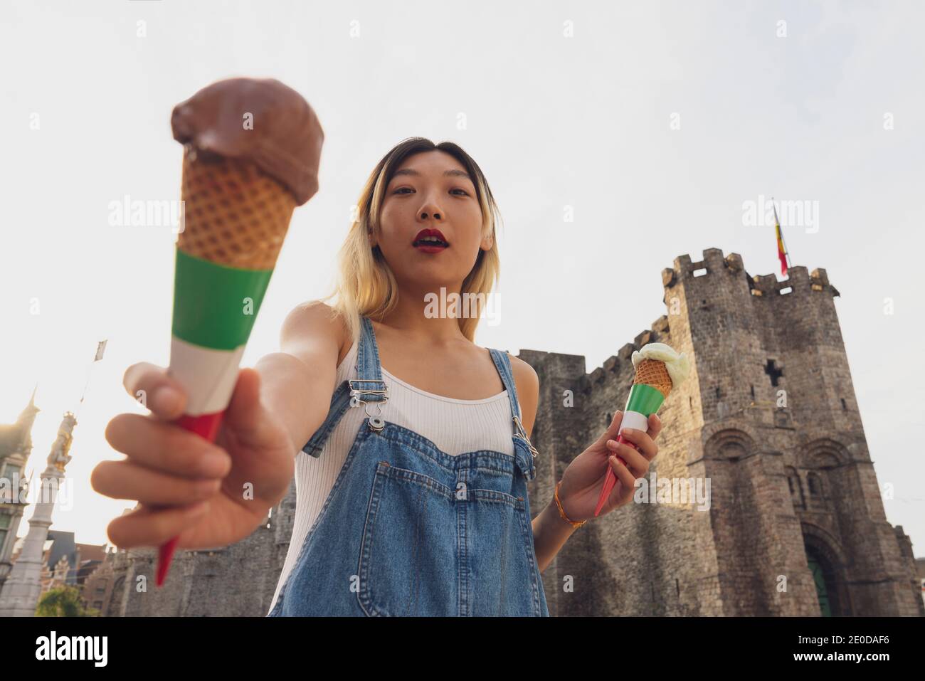 From below of Asian female standing with sweet ice cream in waffle cones in city and looking at camera while enjoying stroll on summer day Stock Photo
