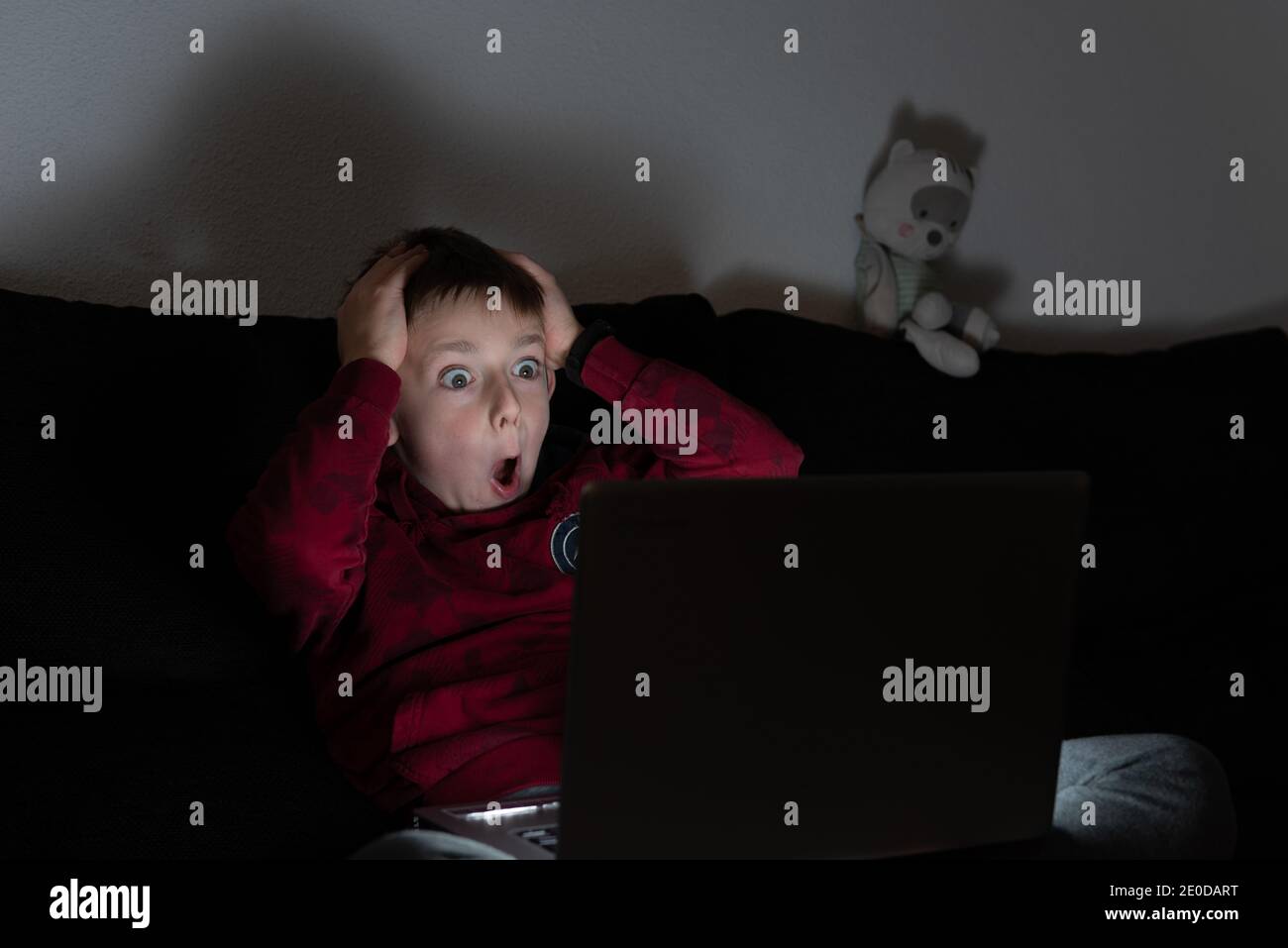 Terrified boy sitting on sofa in dark room and watching horror film on laptop Stock Photo