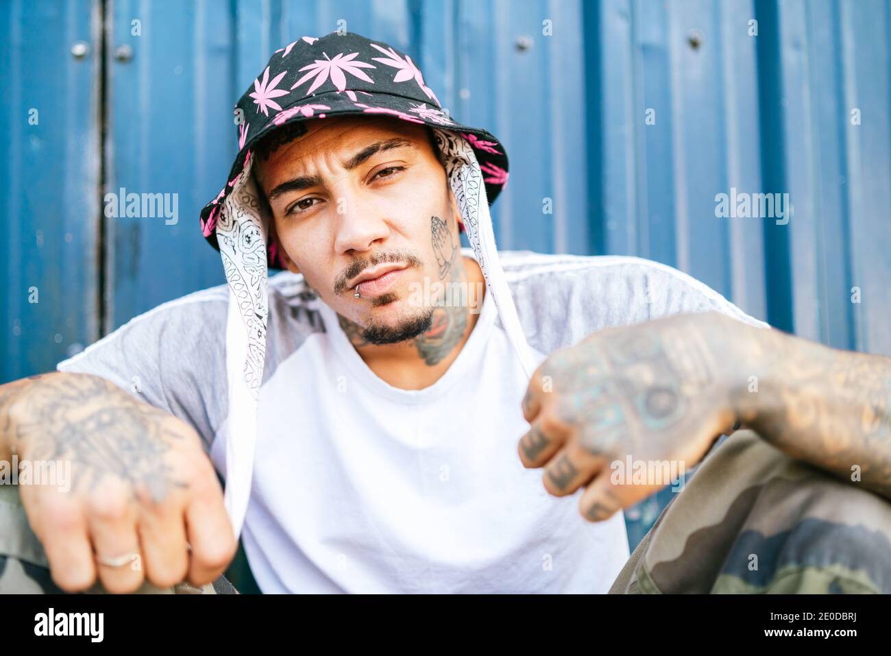 Young tattooed male hipster in informal clothing sitting on ground near metal fence and looking at camera Stock Photo