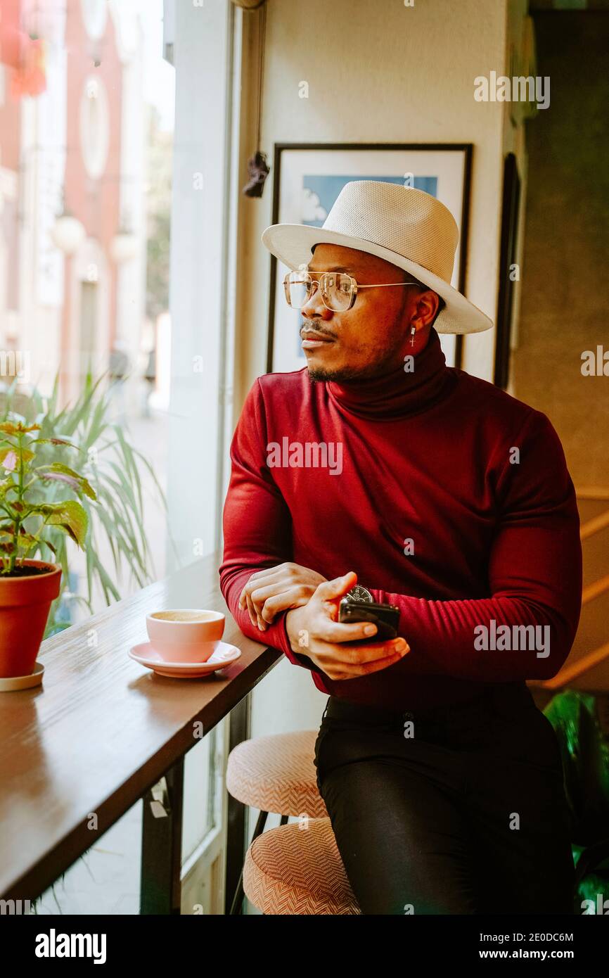 Pensive African American male in stylish outfit sitting at counter in coffee shop while chilling at weekend and looking out of window Stock Photo