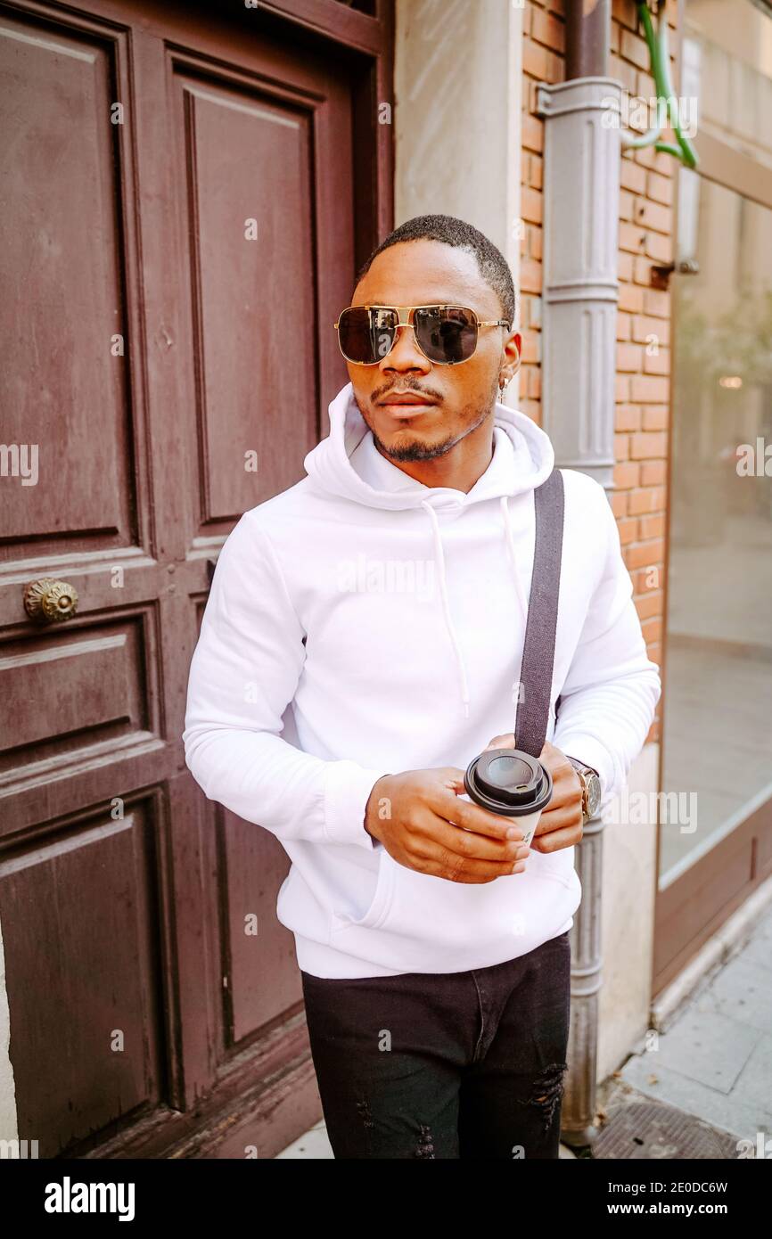 Confident African American male in stylish casual clothes standing on street with coffee to go and looking away during weekend city stroll Stock Photo