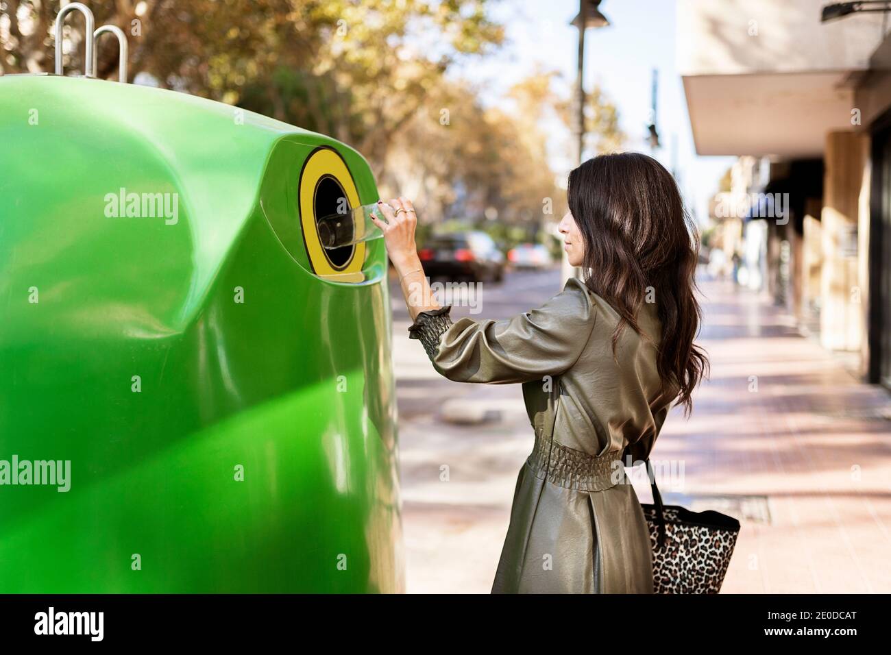 Side view of young stylish ethnic female in leather dress throwing plastic bottle into green recycling bin placed on city street Stock Photo