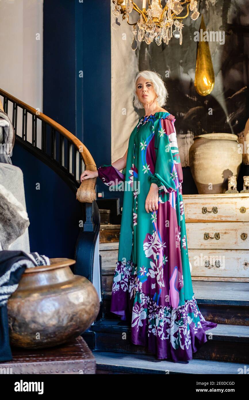 Confident mature female in elegant maxi dress standing near stairs in expensive house while looking at camera Stock Photo