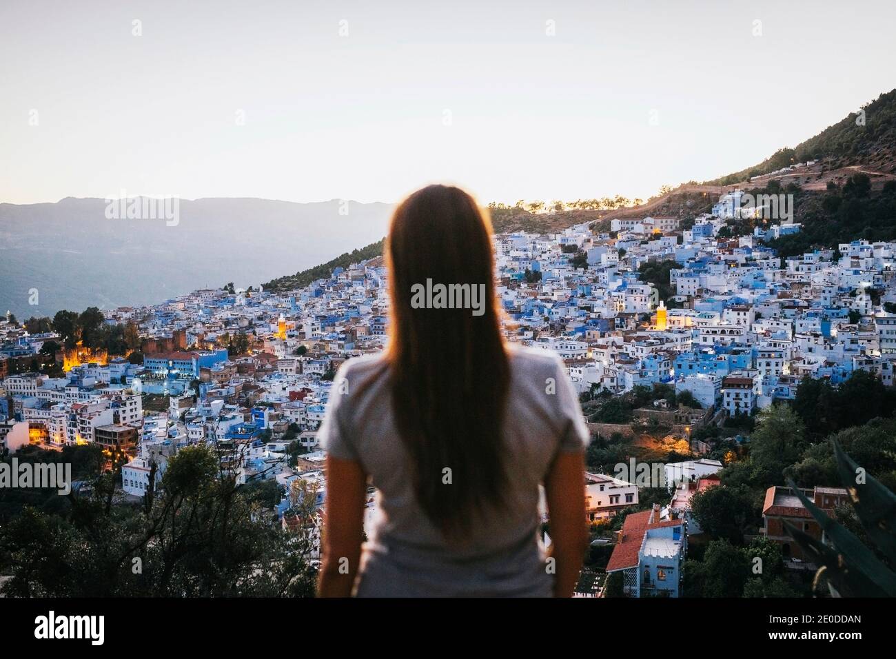 Back view of unrecognizable young female tourist admiring picturesque cityscape with cozy typical white and blue houses located on hill slope against Stock Photo