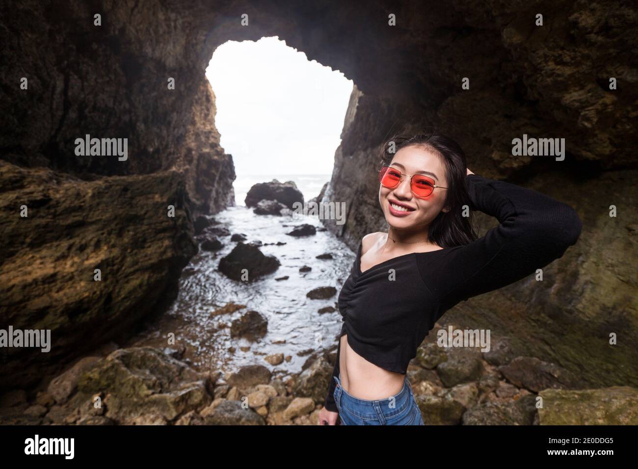 Side view of cheerful young Asian female traveler in casual clothes and sunglasses looking at camera while exploring rocky cave near sea during vacati Stock Photo